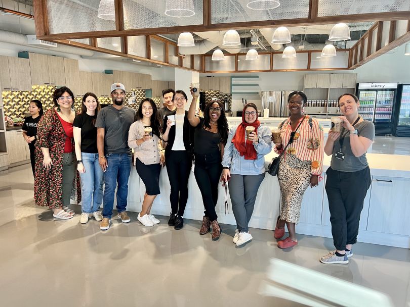 A group of 10 Udemy employees stand next to one another in front of a counter in a large office kitchen, holding coffees and smiling.